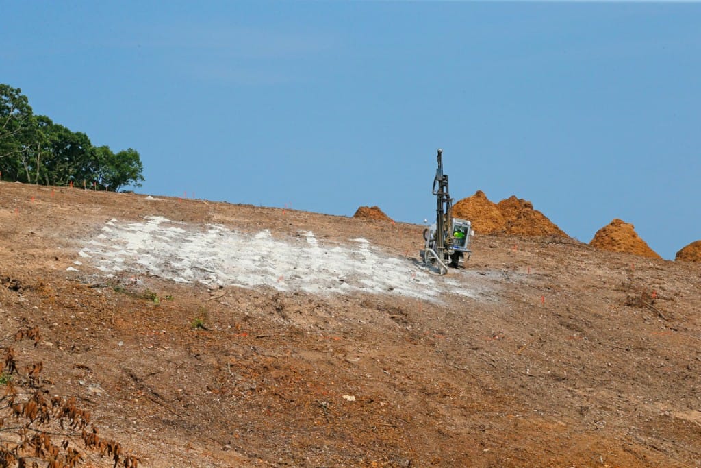A drilling rig from R&K Excavators prepares holes to accept the explosive charges for blasting to move soil and rock down the hillside. (08/22/18)